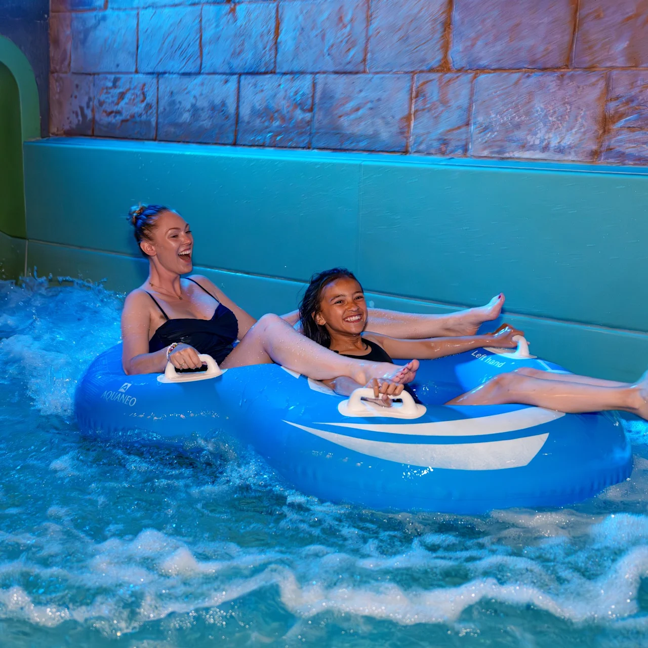 Two people on a blue inflatable tube are enjoying a ride in a water park, laughing and splashing in a pool surrounded by stone walls and blue lighting.