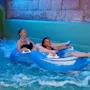To personer på en blå oppustelig badering nyder en tur i en vandland, mens de ler og plasker i en pool omgivet af sten vægge og blåt lys.
