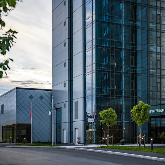En moderne hotellbygning med store glassvinduer reflekterer himmelen, med teksten "QUALITY HOTEL PANO," omgitt av trær og en skyet himmel.