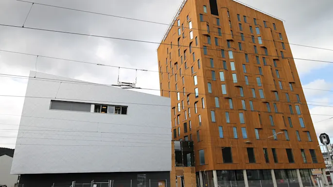 A tall, modern wooden building with irregular windows stands beside a slanted white structure, framed by overcast sky and train tracks. Text: "Mjøstårnet".