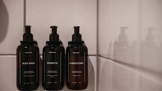 Tre mörka pumpflaskor märkta Body Wash, Schampo och Balsam i en hotell dusch.