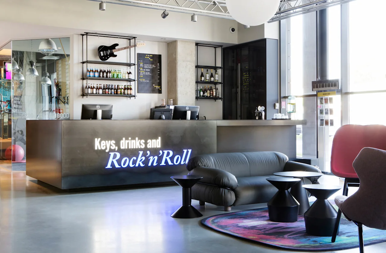A modern reception desk displays the text "Keys, drinks and Rock'n'Roll" with a guitar and bottles above. Comfortable seating and colorful decor enhance the welcoming lounge atmosphere.