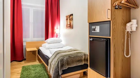 Et lite, moderne hotellrom har en enkeltseng med hvitt sengetøy og et brunt teppe, ved siden av røde gardiner. Et skap med safe og minikjøleskap står i nærheten.
