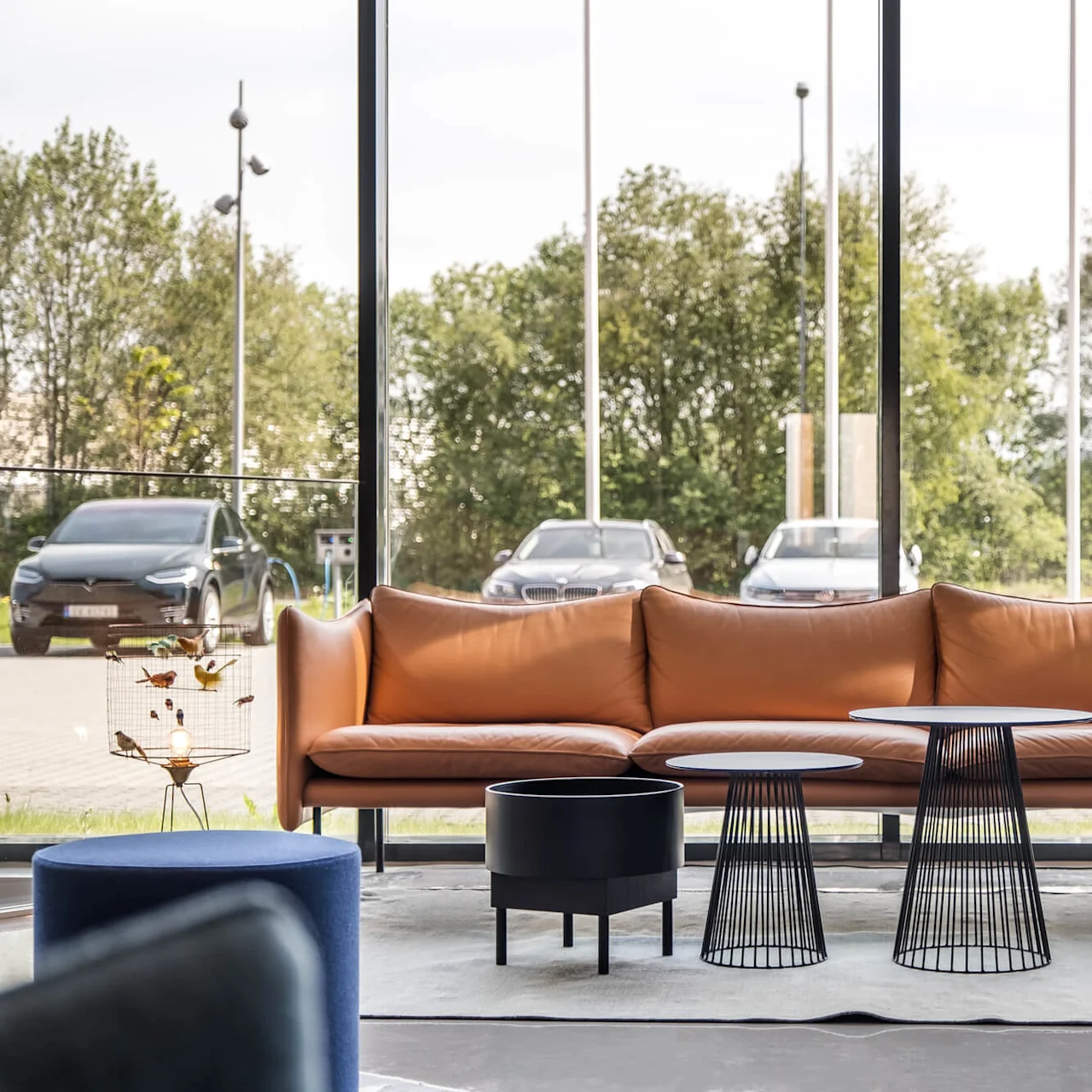 Brown couch sitting in a modern lobby near two black wire-frame tables; visible through large windows, parked cars and trees line a sunny parking lot.