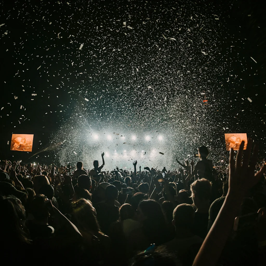 A large crowd at a lively concert with confetti falling, hands raised in excitement, and bright stage lights. Signs show 2 and 3.