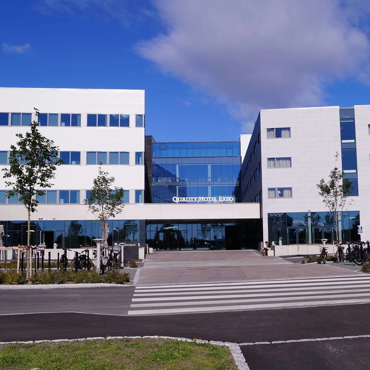 A modern hotel building labeled "Quality Hotel Expo" stands under a blue sky. Bicycles are parked in front, with young trees lining the entrance.