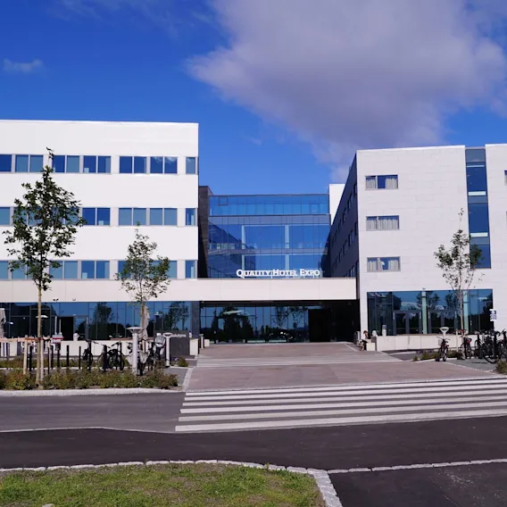 En moderne hotellbygning med teksten "Quality Hotel Expo" står under en blå himmel. Sykler er parkert foran, med unge trær langs inngangen.