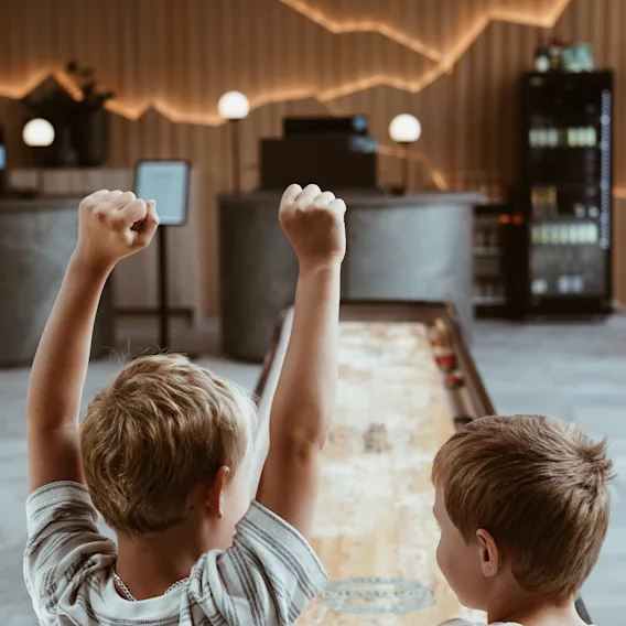 Barn spelar shuffleboard på Quality Hotel Arlanda XPO. Ett barn firar en poäng. Synlig text: BEWARE The game consists of nutshells.