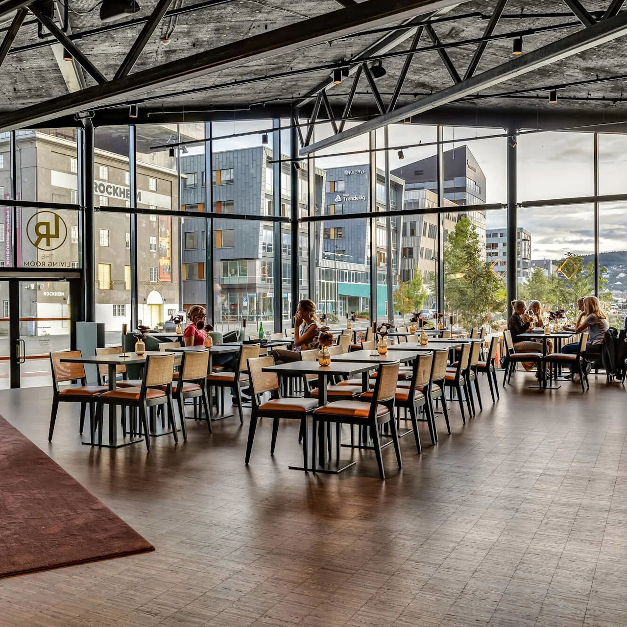 Restaurant interior with modern decor; people sit at tables, enjoying drinks near large windows showcasing an urban cityscape. Text present: "THE LIVING ROOM," "ROCKHEIM," "Trøndelag."