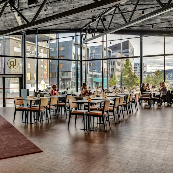 Restaurantinteriør med moderne innredning; mennesker sitter ved bord og nyter drikker nær store vinduer som viser et bylandskap. Tekst til stede: "THE LIVING ROOM," "ROCKHEIM," "Trøndelag."