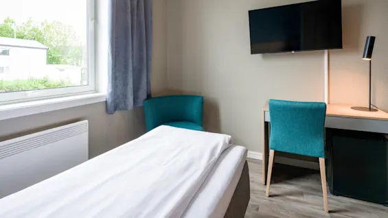Bed with white sheets in a small hotel room, positioned near a window. A desk with a chair, lamp, and TV on the wall occupies the corner.