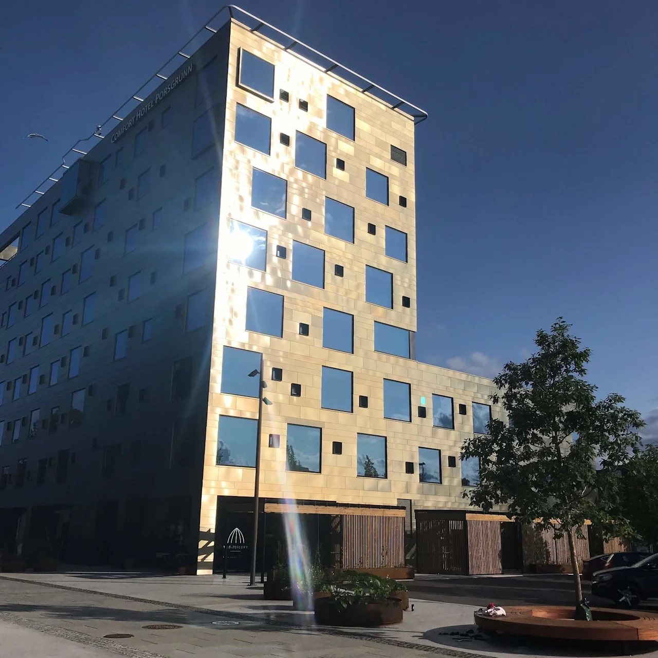En moderne, guldfarvet bygning med firkantede vinduer reflekterer sollys. Et træ og gadeinventar foran. Tekst: "Comfort Hotel Porsgrunn."