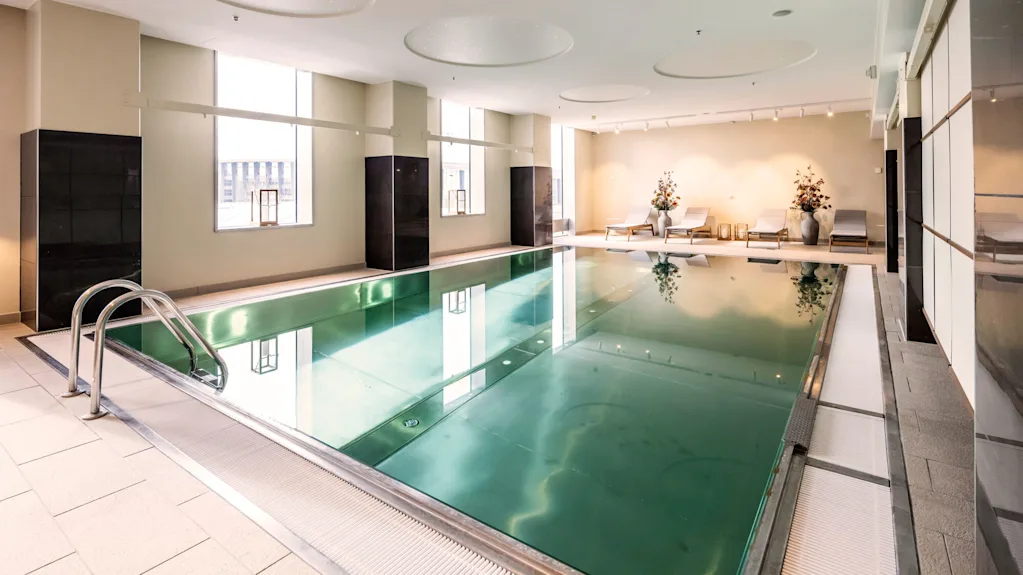 Swimming pool indoors, reflecting large windows on the left. Lounge chairs and decorative plants placed on the right side. Bright ceiling lights illuminate the modern, minimalist space.