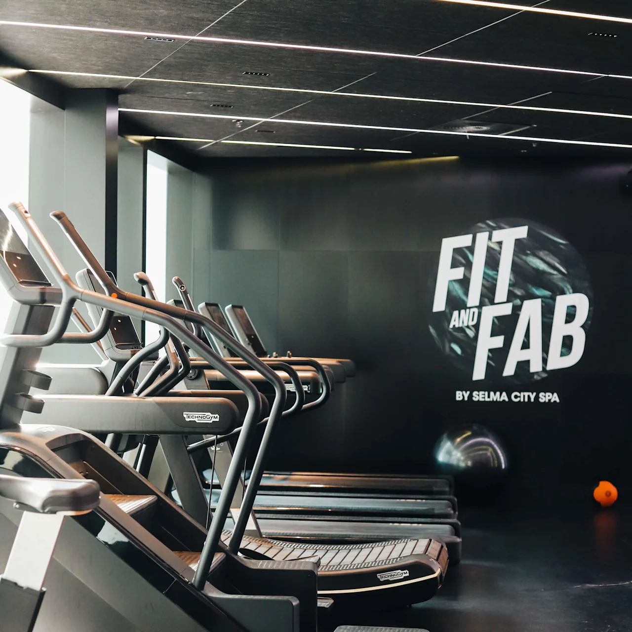 Löpband står på rad i ett modernt gym med stilrena mörka interiörer. På en vägg står texten "FIT AND FAB BY SELMA CITY SPA." En orange träningsboll ligger på golvet.