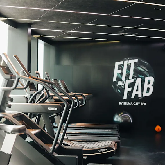 Löpband står på rad i ett modernt gym med stilrena mörka interiörer. På en vägg står texten "FIT AND FAB BY SELMA CITY SPA." En orange träningsboll ligger på golvet.