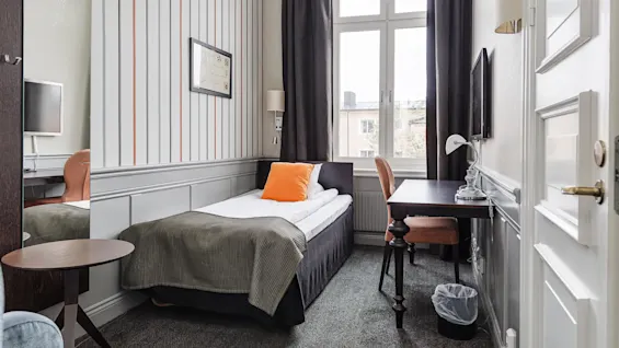 A neatly arranged small bedroom with a single bed, a desk, chair, window, and striped wallpaper.