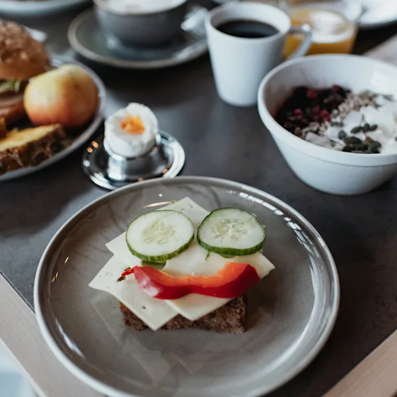 Öppen smörgås toppad med ost, gurka och paprika på en tallrik; omgiven av frukostartiklar som kokt ägg, kaffe, frukt och en skål med müsli i matsal.