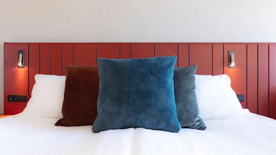 A neatly made bed with plush pillows, highlighted by a warm wall lamp, against a red paneled wall.