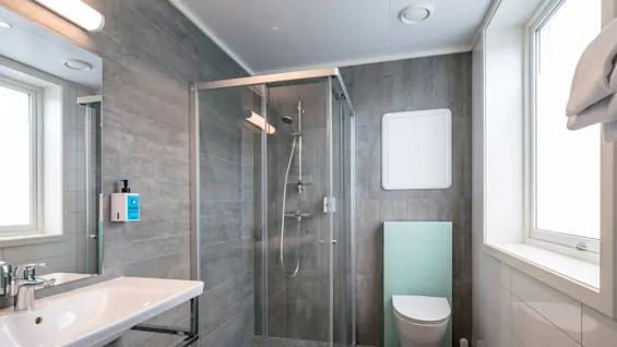 A glass shower enclosure stands unused in a modern bathroom with grey tiles, adjacent to a wall-mounted sink and toilet, illuminated by natural light from a window.