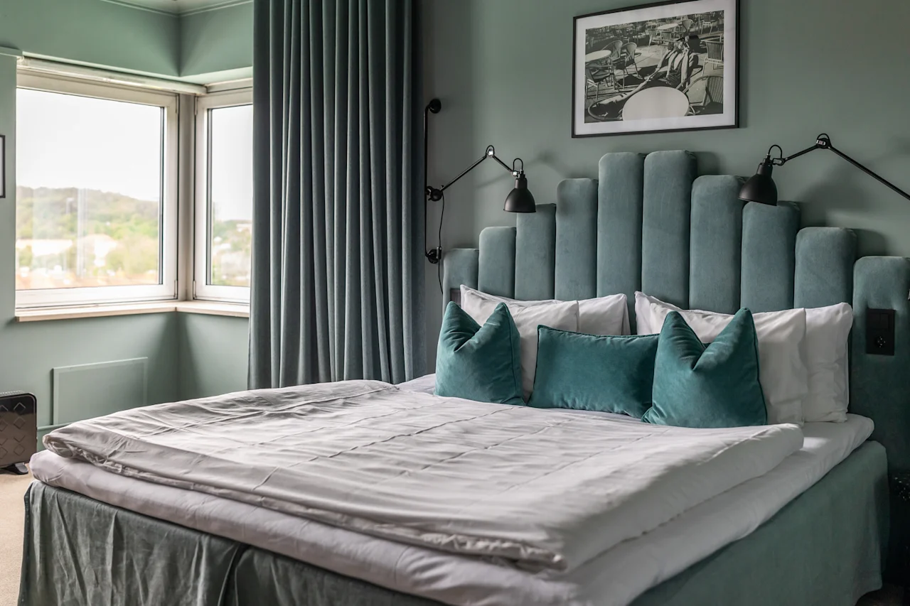 Mukava hotellihuone, jossa on muhkea sänky turkoosilla sängynpäädyllä, pehmeät vuodevaatteet ja valoisa ikkunanäkymä.