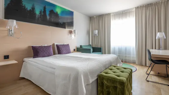 En säng med vita lakan och lila kuddar står bredvid en träpanel i ett ljust hotellrum. En grön ottoman, skrivbord och stolar kompletterar den moderna inredningen.
