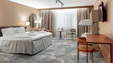 Ett prydligt arrangerat hotellrum har en stor säng med vita sängkläder, en våningssäng, ett skrivbord med lampa, ett bord med stol och mjukt ljus genom gardiner.