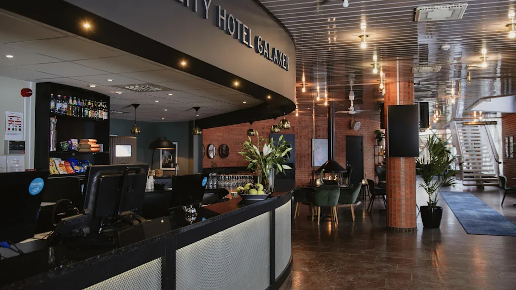 Receptionen visar texten "QUALITY HOTEL GALAXEN" med datorer och snacks. Tegelväggar, gröna stolar och bord i ett varmt belyst lobby. Trappa och växter förhöjer den moderna känslan.