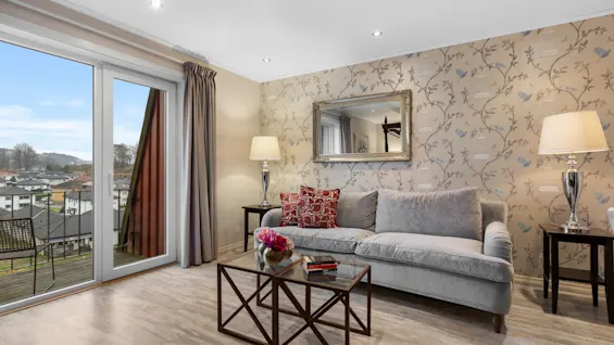 Et koselig oppholdsrom med en grå sofa, salongbord og lamper, med en balkong som vender ut mot en by og åser.