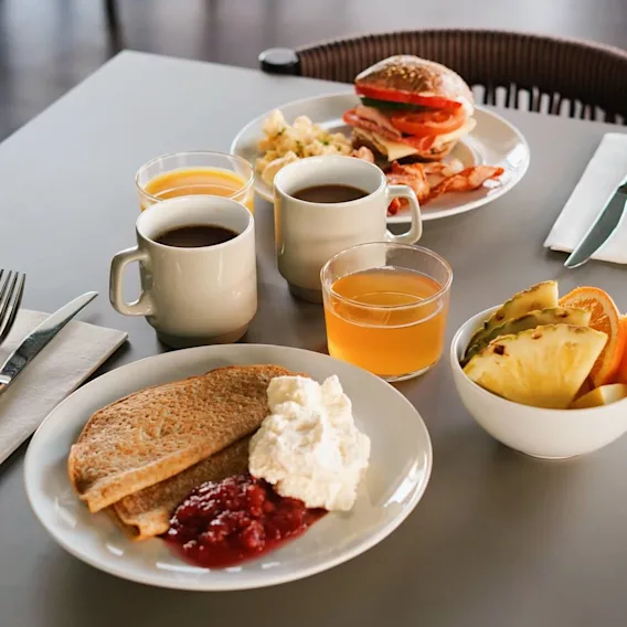Tallrikar med frukostmat, inklusive pannkakor, frukt och en smörgås, står på ett bord med juice och kaffe i en restaurangmiljö.
