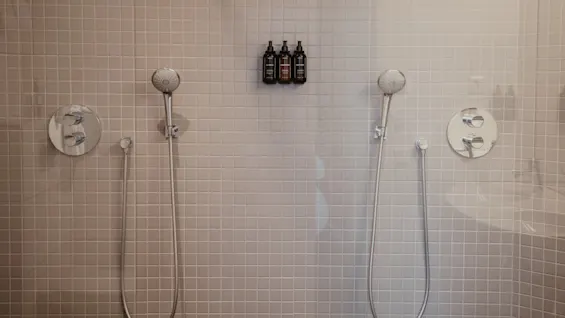 To brusehoveder hænger fra en flisebelagt badeværelsesvæg, med tre sorte flasker monteret centralt. Indstillingen er et moderne, minimalistisk badeværelse med neutrale toner.