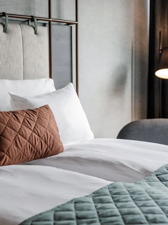 En bekväm hotellsäng med vita kuddar, en brun prydnadskudde och en turkos filt, redo för en avkopplande vistelse.
