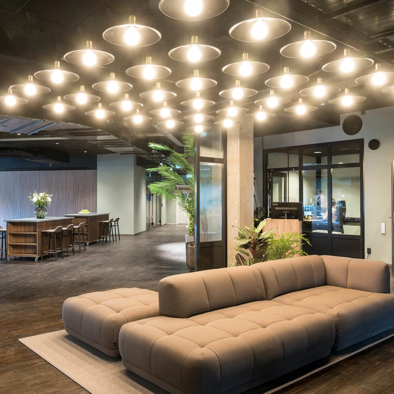 A spacious, modern lounge features a brown sofa under bright ceiling lights. Nearby, a tall table with stools and plants create a welcoming atmosphere in a sleek office setting.