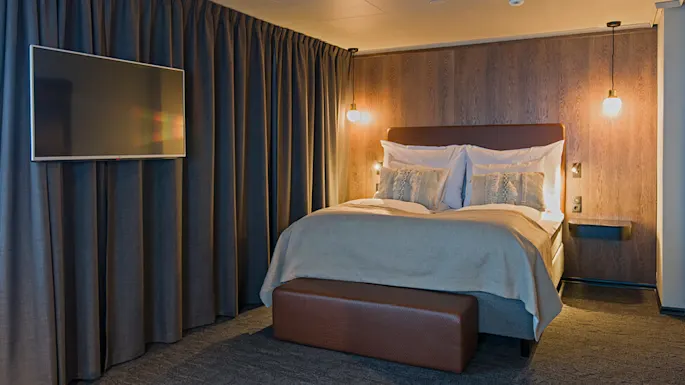 A large bed with pillows and a blanket is centered against a wooden wall, illuminated by hanging lamps. A television is mounted on the left, with curtains in the background.