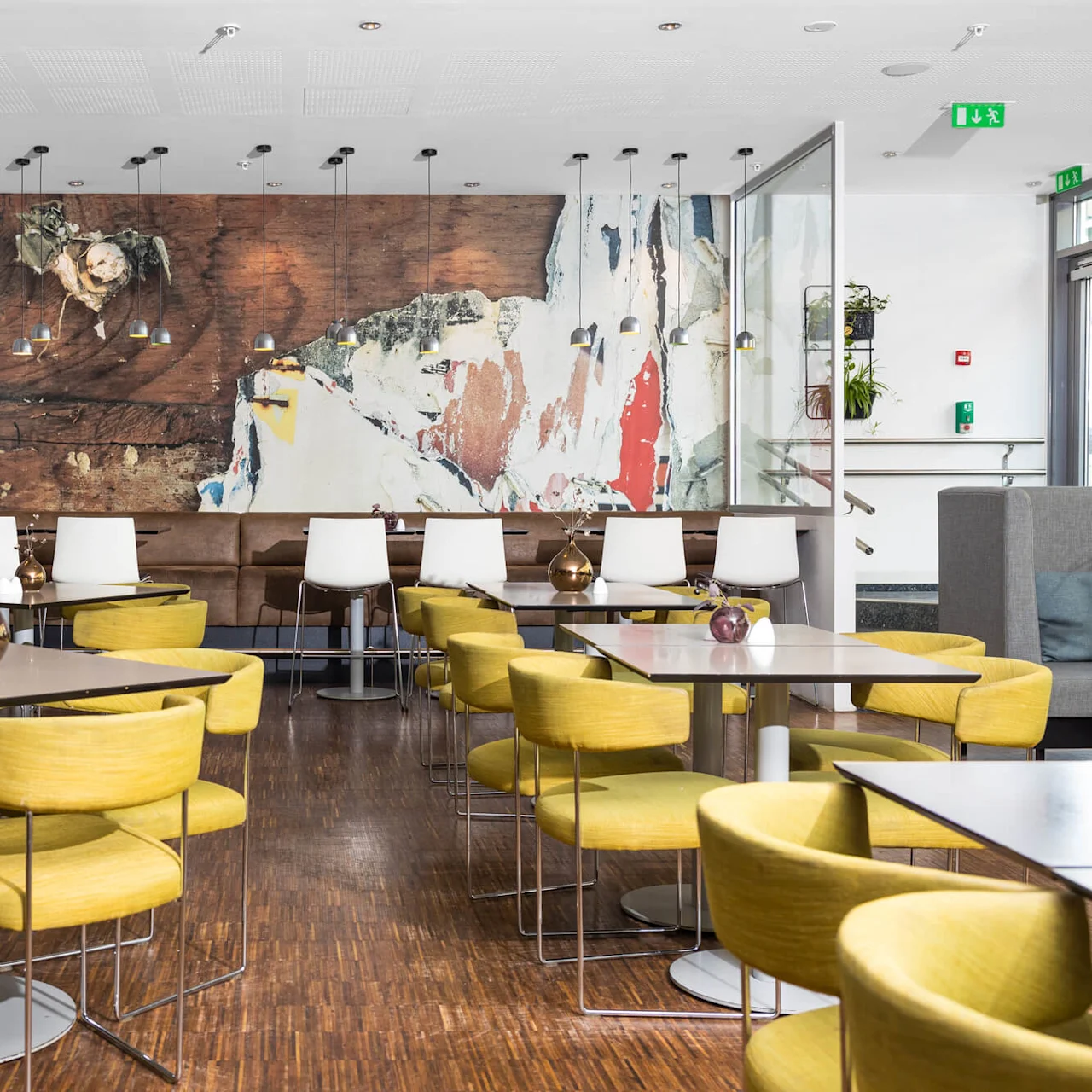 Tables with yellow chairs are arranged in a modern café. A colorful abstract mural decorates the wall. Overhead lights and modern decor create a stylish atmosphere.
