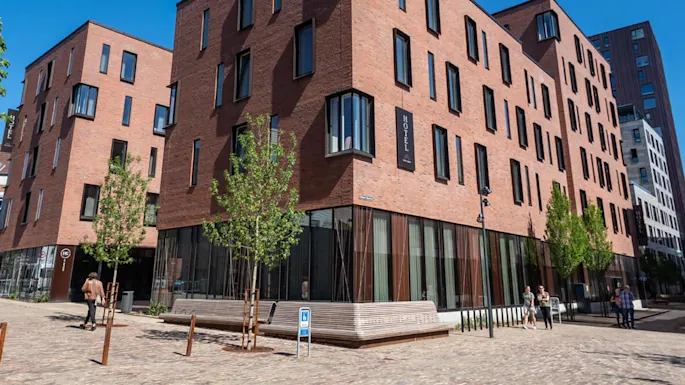 En moderne murbygning i flere etasjer som er et hotell med store vinduer ligger i en gate med trær og benker. Folk går i nærheten. Tekst: "HOTEL".