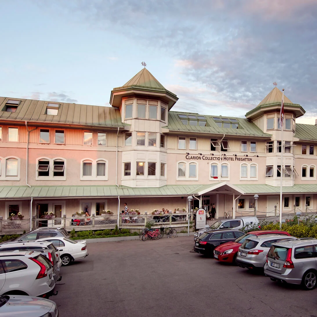 Ett historiskt hotell med en terrass och gäster vid skymning, omgivet av parkerade bilar.