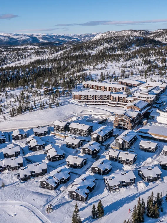 Snow-covered village nestled in mountainous terrain, surrounded by evergreen trees. The residential complex consists of multiple buildings. Ski tracks and a distant horizon of wooded hills complete the winter scene.