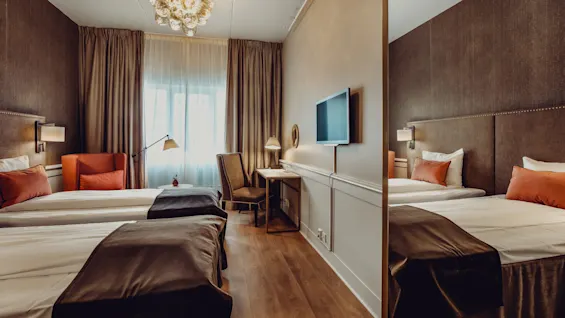A twin hotel room with two beds, a desk, TV, and chair, softly lit with an elegant, cozy decor.