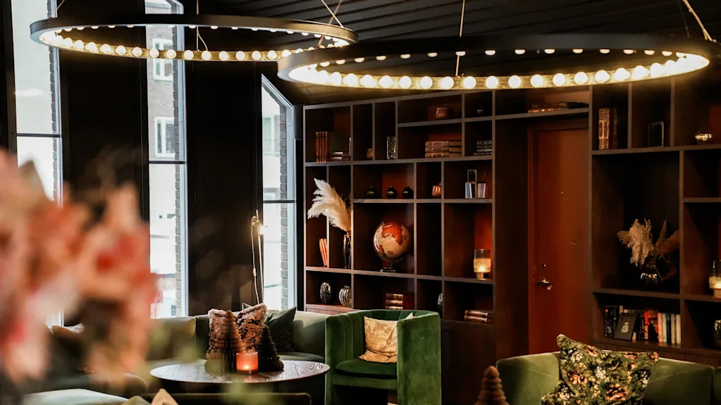 Elegant lounge with plush green chairs surrounds wooden tables, warmly lit by circular ceiling lamps. Shelves display books, a globe, and decorative items, adding a cozy atmosphere.