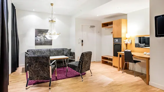 En stue har en krystallysekrone over en sortmønstret sofa med et lite kaffebord på et lilla teppe. Et skrivebord og hylle i tre opptar høyre side.
