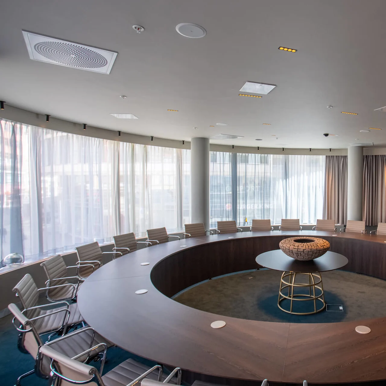 A large, circular conference table is surrounded by office chairs, situated in a room with floor-to-ceiling curtains and a big screen on the wall.