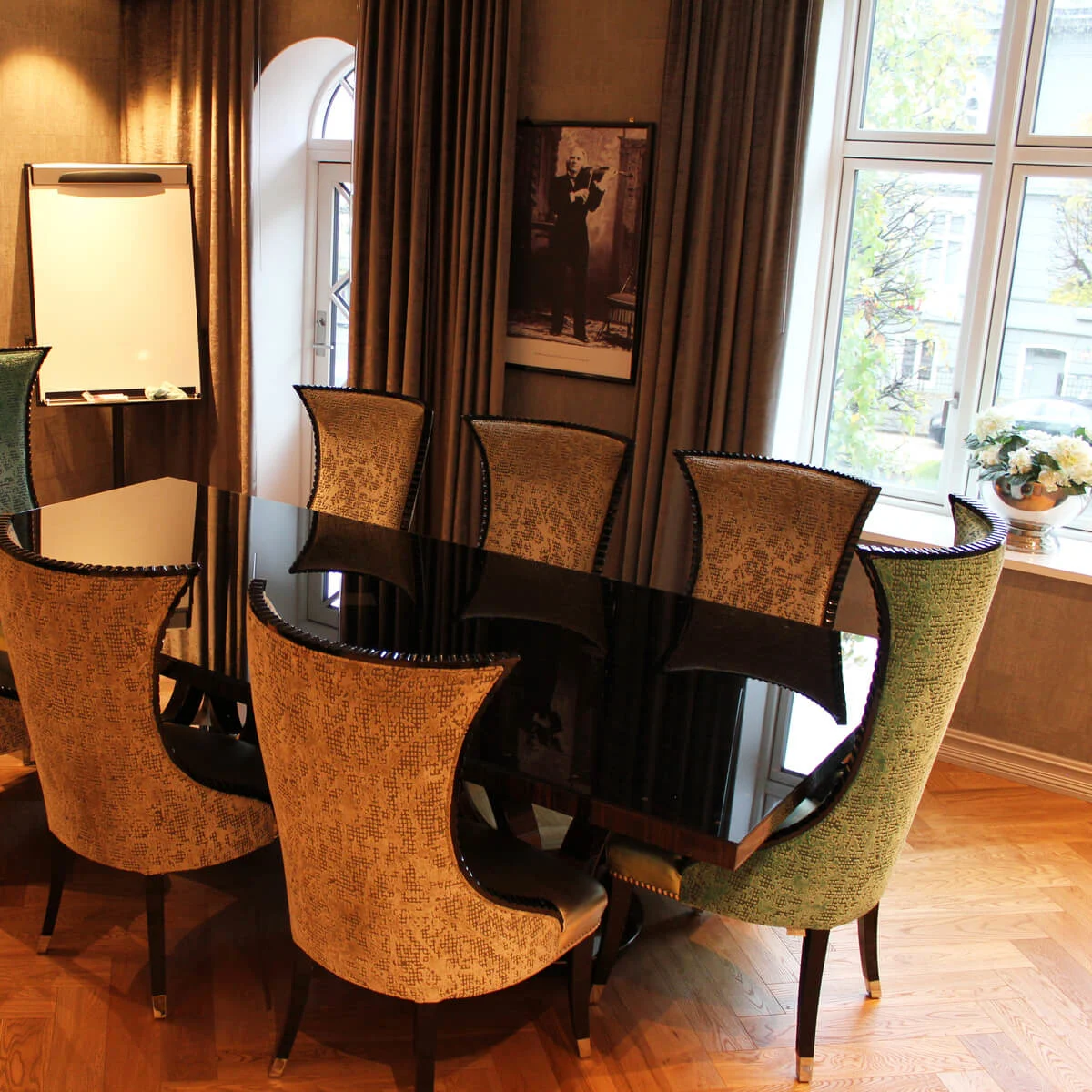 A black glass table surrounded by elegant chairs stands in a well-lit room, featuring a TV, whiteboard, and large windows with curtains.