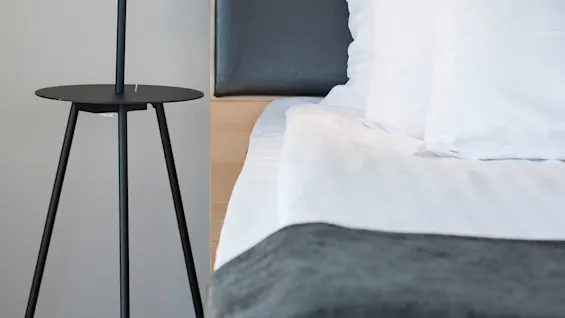 A black floor lamp stands on a modern bedside table, illuminating a neatly made bed with white pillows and sheets, set against a dark headboard.