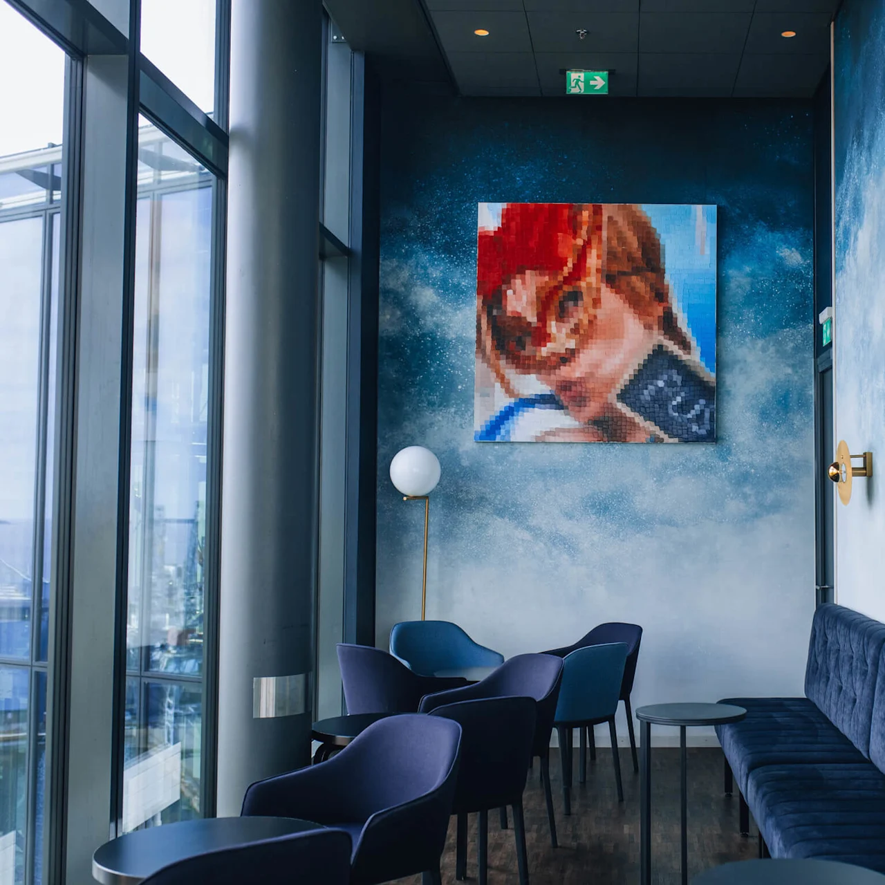 Elegante stole og borde er arrangeret i en moderne lounge med strukturerede blå vægge, blød belysning og et farverigt mosaik kunstværk. Store vinduer giver naturligt lys og udsigt udenfor.
