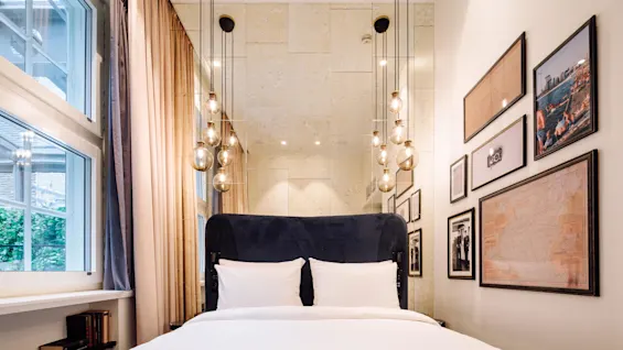 A bed with white sheets is positioned against a wall adorned with framed pictures. Hanging lights dangle above, while large windows and curtains are on the left.