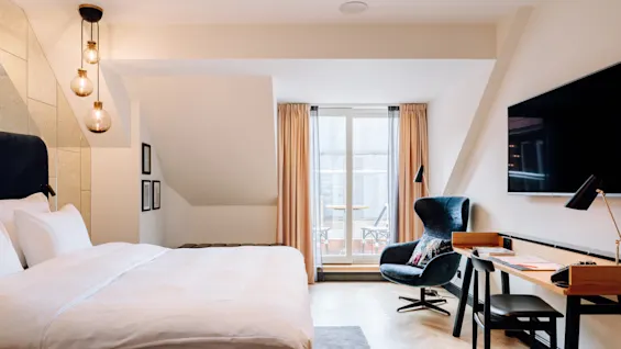 A well-lit hotel room contains a bed with white linens, a desk with a lamp and chair, and an armchair near a window with sheer curtains. 