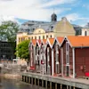 Red wooden warehouses line a calm river, bordered by historical brick and stucco buildings under a partly cloudy sky. A stone bridge, lush trees, and a bustling city scene complete the view.