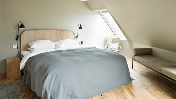 Bed with gray bedding stands in a minimalist room. Wall lamps are mounted above nightstands; a wicker chair sits by a slanted window. Wooden floor complements soft colors.