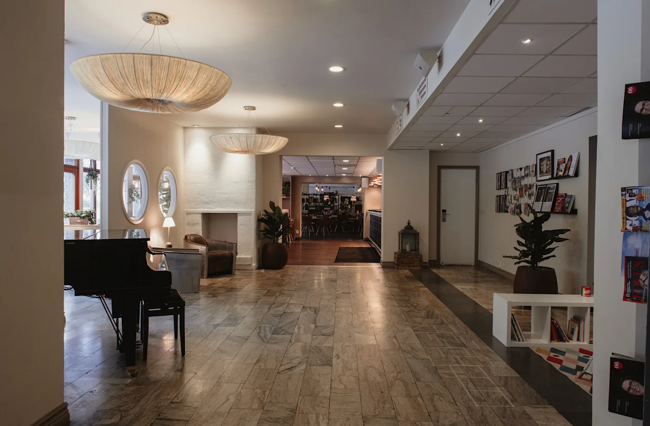 A spacious and inviting hotel lobby with a grand piano, comfortable seating, and a view into a bustling restaurant area.
