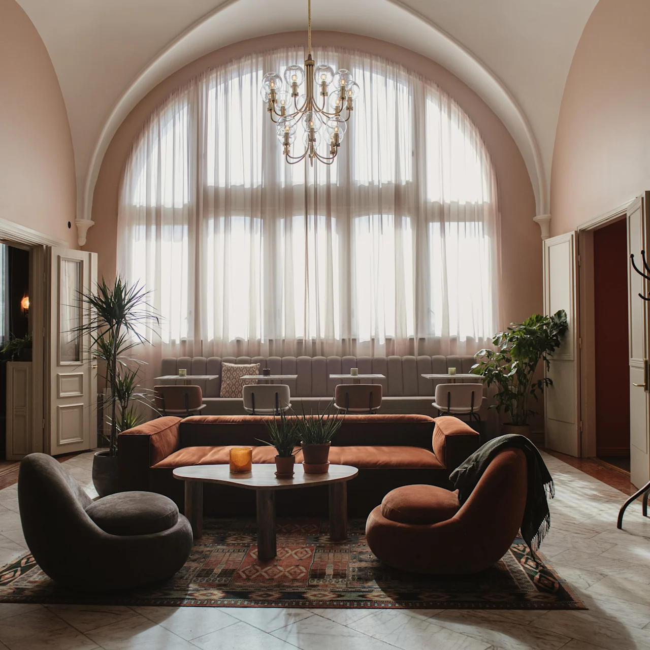 Mukava lounge-tila sisältää pyöreän sohvapöydän, jota ympäröivät modernit tuolit ja sohva. Aurinko paistaa suurista kaari-ikkunoista korostaen kasveja elegantissa huoneessa.
